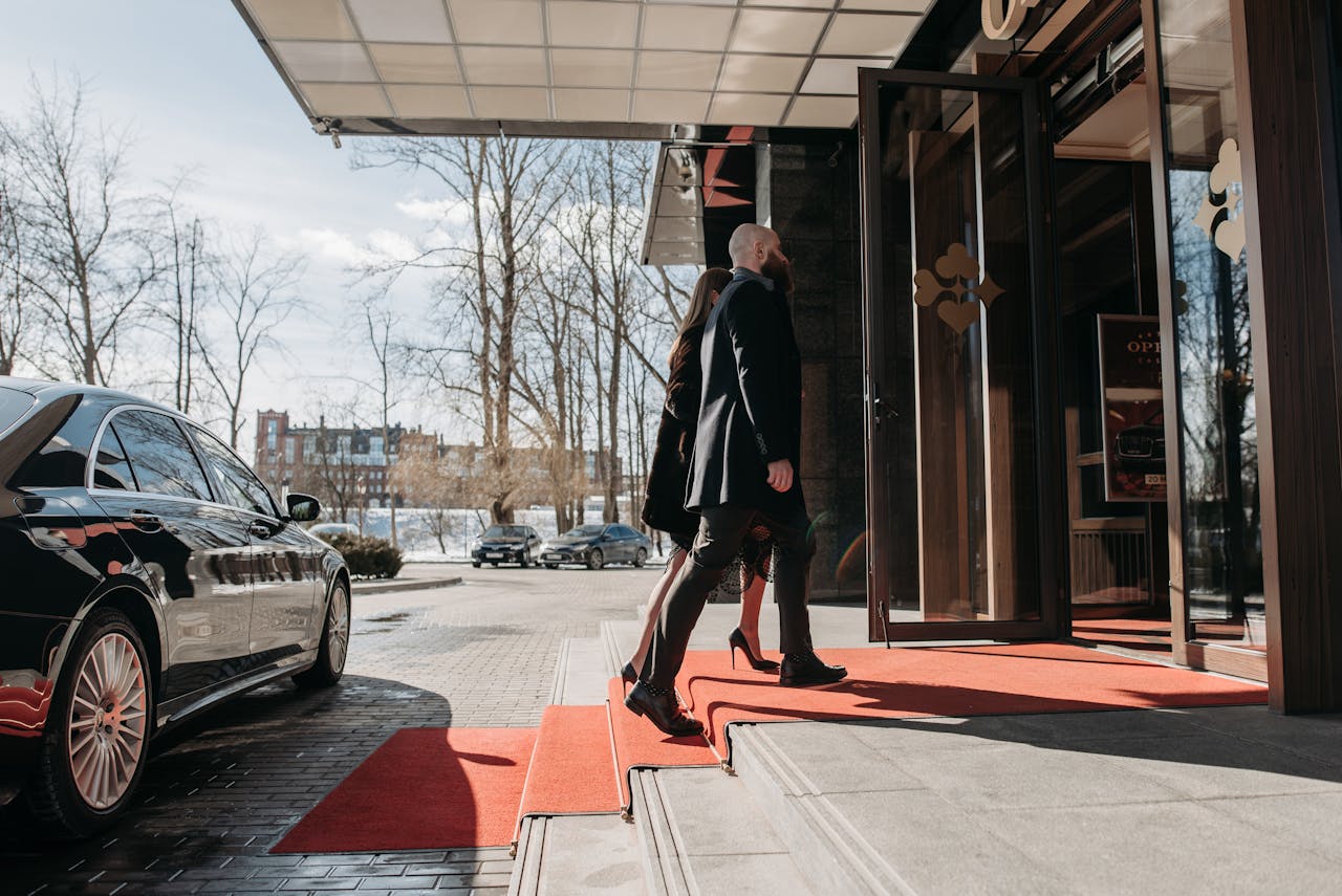 gallery-img-03 A man and woman elegantly dressed enter a luxury hotel, walking on a red carpet.